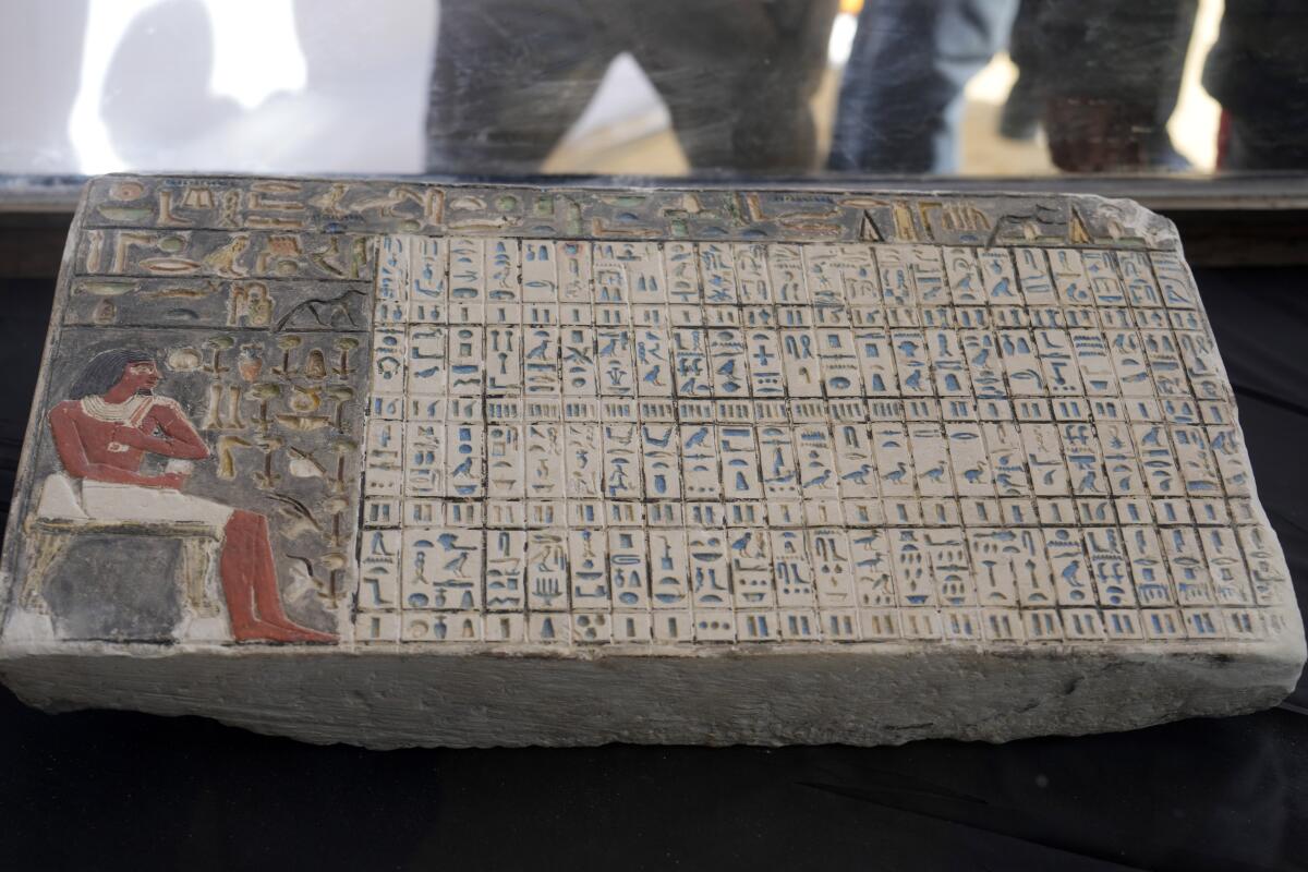 A stone artifact depicts a seated person on the left looking at rows of hieroglyphic symbols 