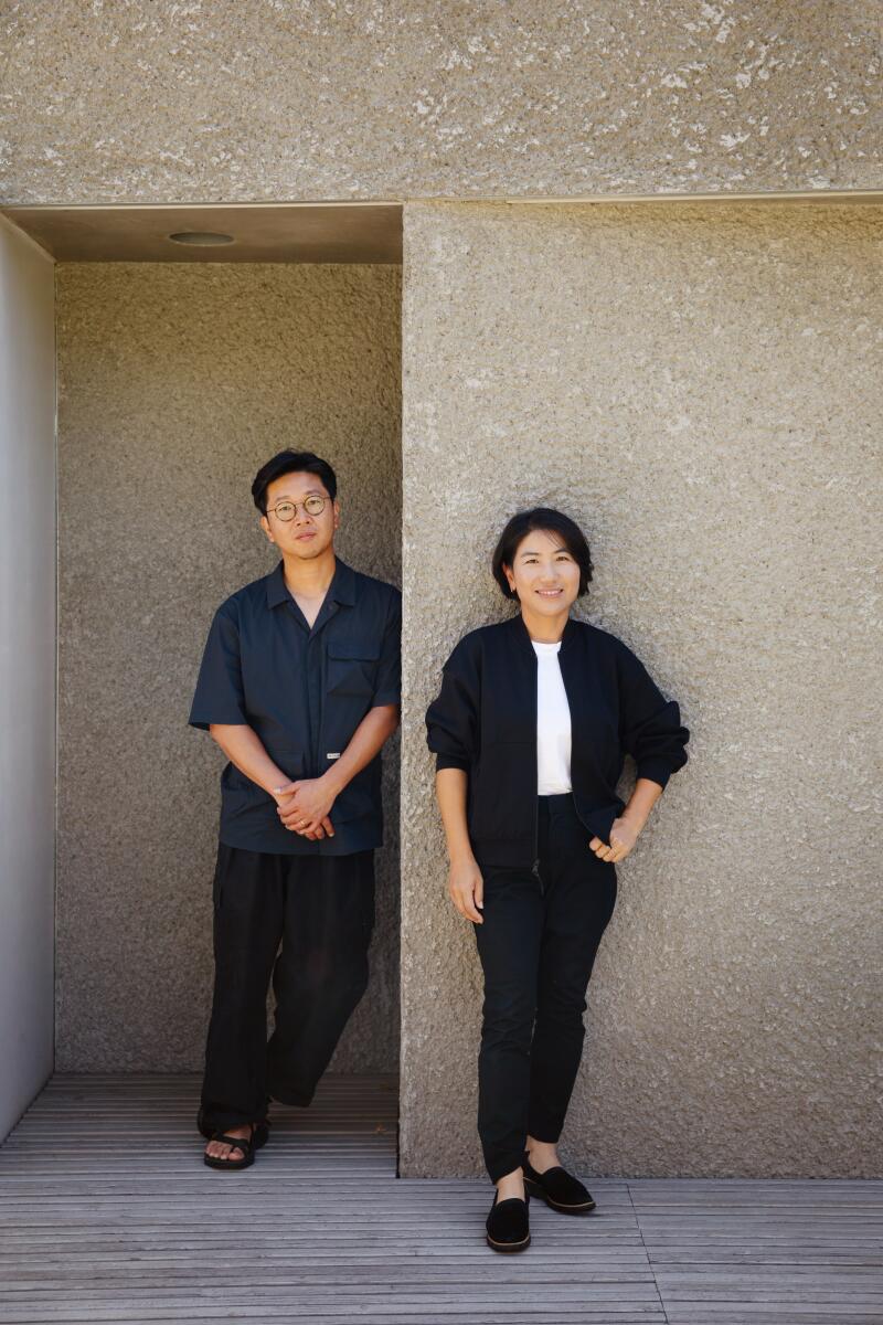 A man and a woman in dark clothing stand against a gray wall