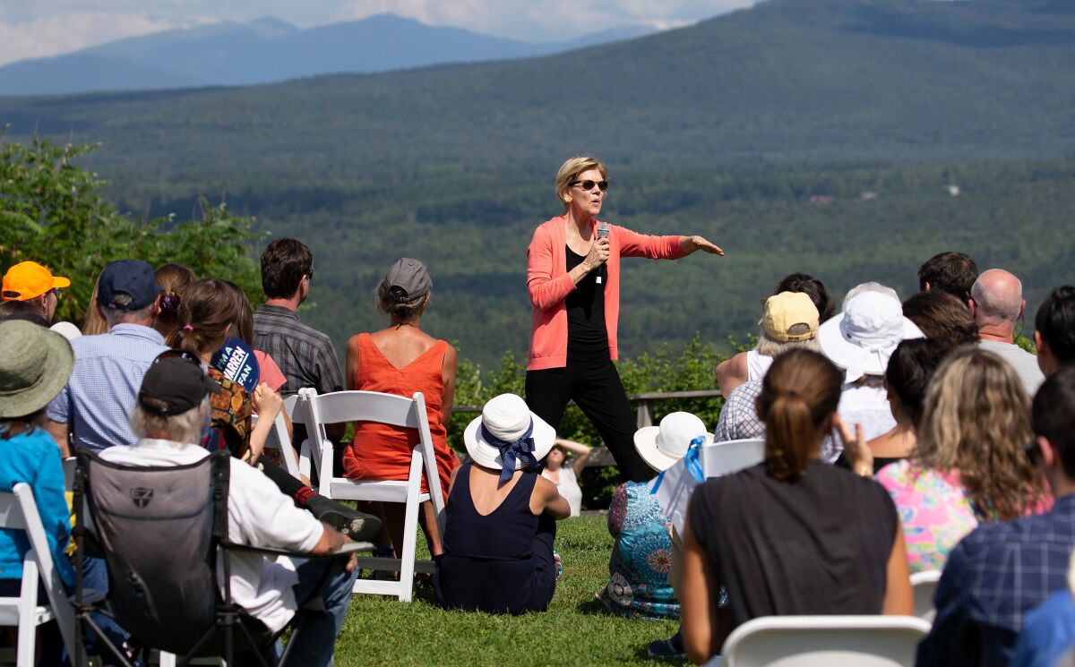 Sen. Elizabeth Warren standing in a field with a microphone speaking to a crowd seated in front of her