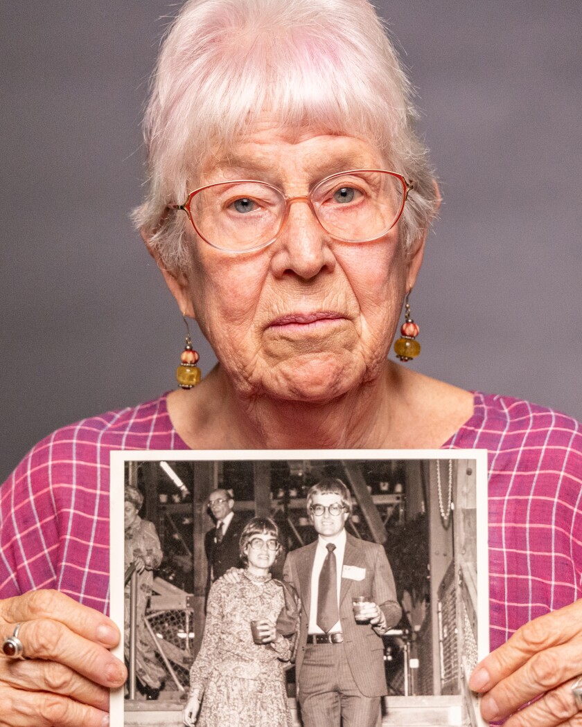 A portrait of a woman wearing a pink shirt holding up a black-and-white photo of a young couple