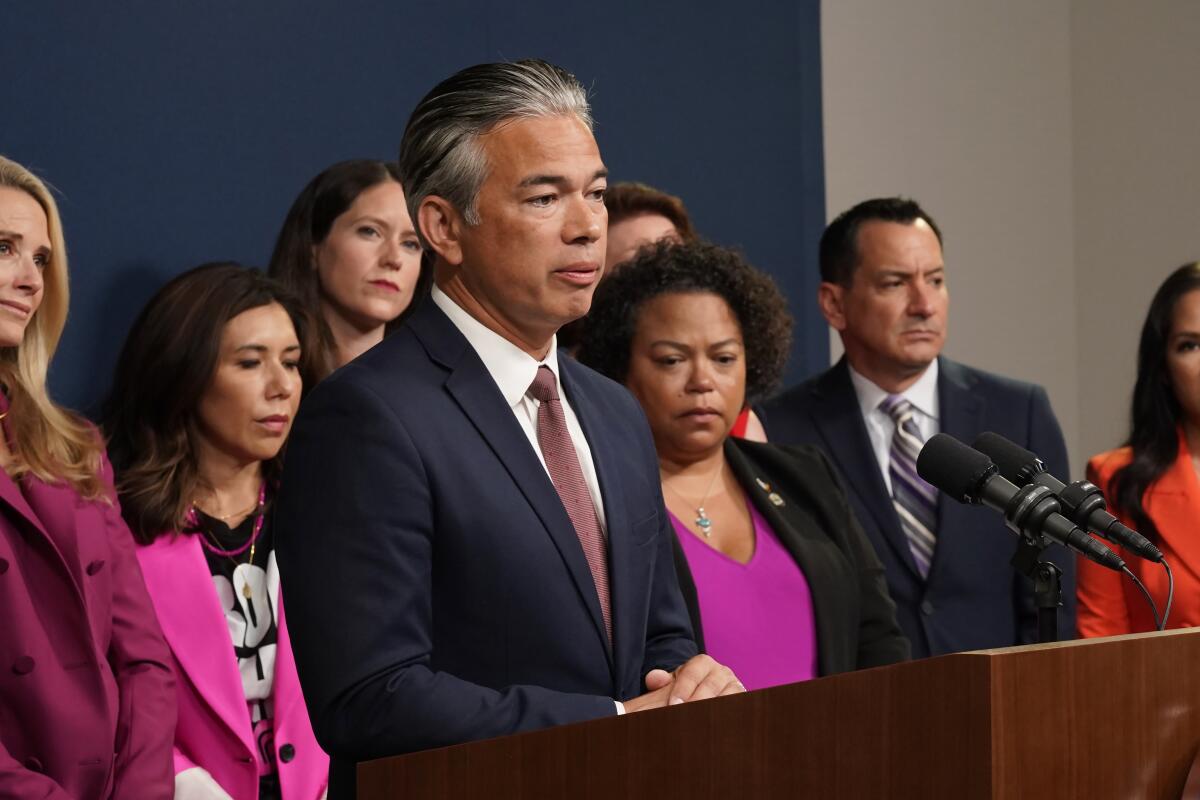 A man in a suit stands at a lectern with several women and one man behind him.