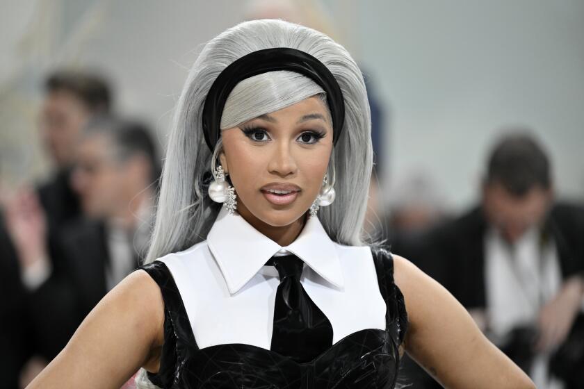 Cardi B poses in a long gray wig, black headband, dangly earrings and a black-and-white suit and tie ensemble.