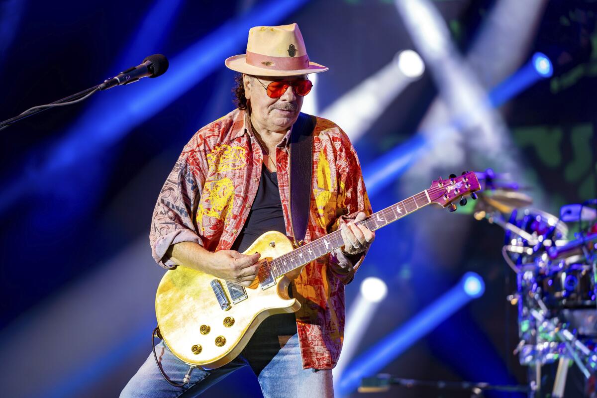 Carlos Santana wears an orange/yellow shirt with a black undershirt and blue jeans as he plays a gold guitar onstage