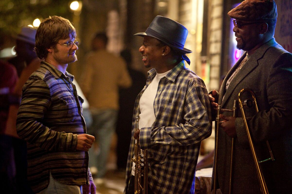 A man holding a trumpet and a man holding a trombone talk to a man wearing glasses.