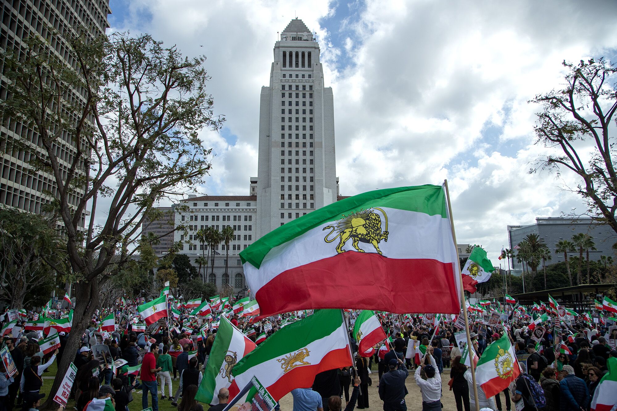 People, many carrying Iran's historic flag, gather in an open area near the City Hall tower