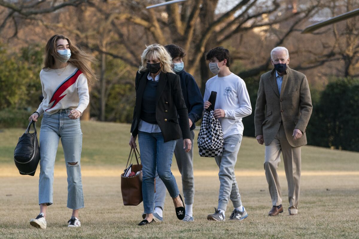 Five people, some carrying bags, walk across a lawn, with trees in the background
