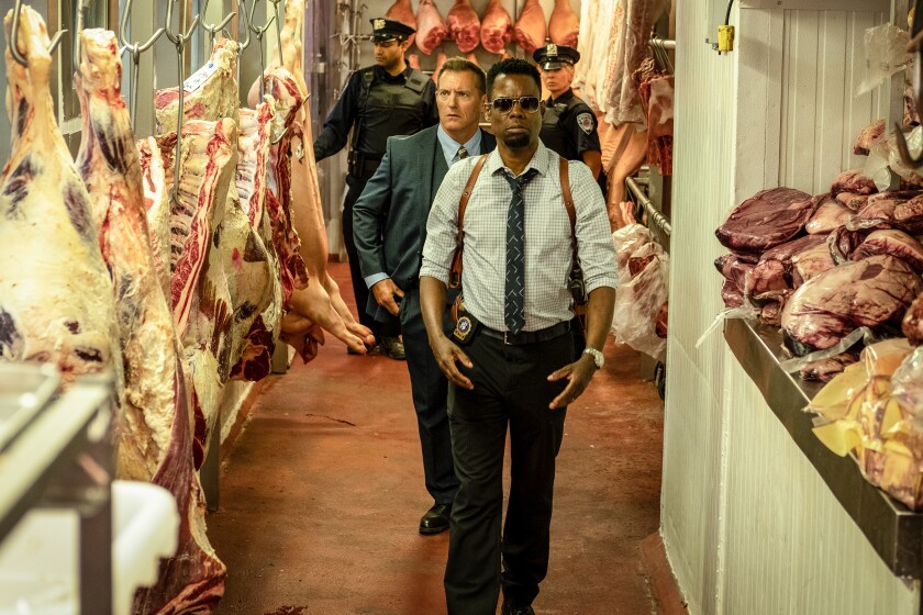 Chris Rock, followed by a man in a suit and two uniformed police officers, walk past hanging meat in a slaughterhouse.