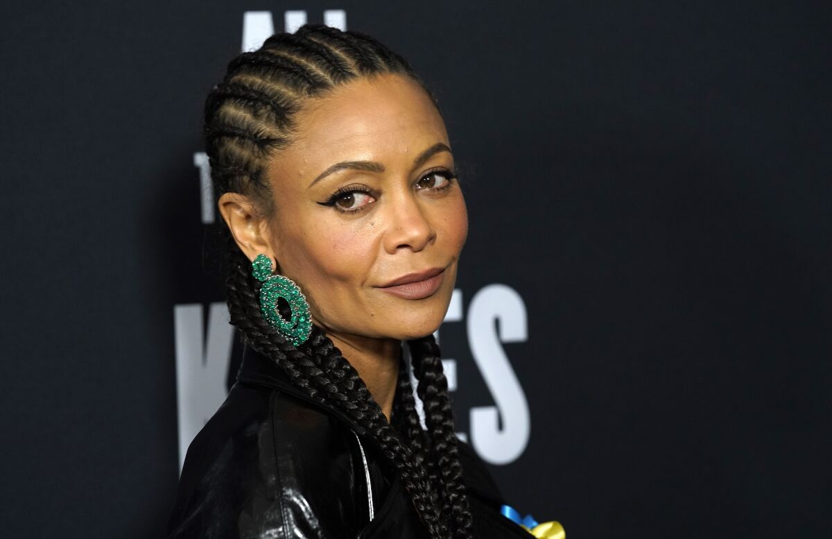A woman with braided hair and large green earrings looks out of the corner of her eye.