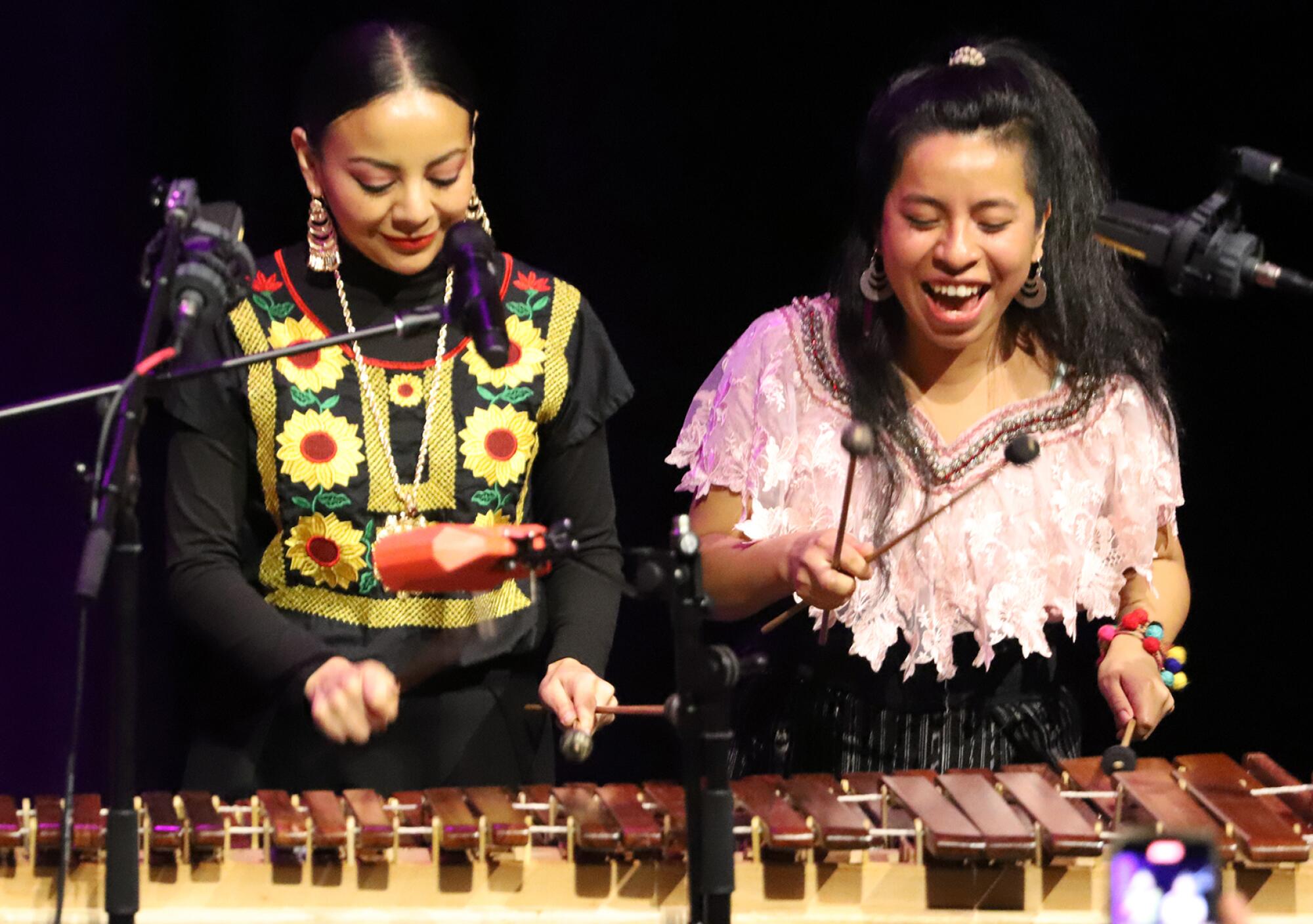 Two women, one dressed in a shirt with yellow-and-red flowers, hold sticks as they play a xylophone-like instrument