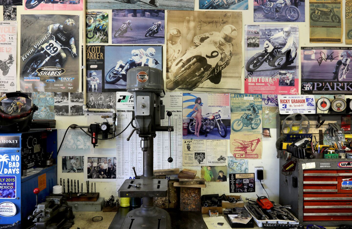 View of a wall in the garage workshop of motorcycle frame builder Jeff Cole showing a drill press, tool box and photos and posters of prominent motorcycle racers from the past on bikes he designed.