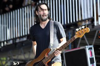 Keanu Reeves, hair long and unshaven, holds bass guitar while wearing a black t-shirt, performing on stage with Dogstar.