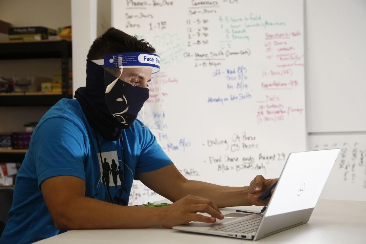 Wearing a face covering and face shield while working amid the coronavirus, Maico Olivares, lead voter registration organizer for Central Arizonans for a Sustainable Economy, works his phone and computer as he tries to reach about 25 people a day, mostly within the Latino community, to persuade them to register to vote Thursday, Aug. 6, 2020, in Phoenix. Like others who register people to vote, those efforts have become extremely difficult during the pandemic. (AP Photo/Ross D. Franklin)