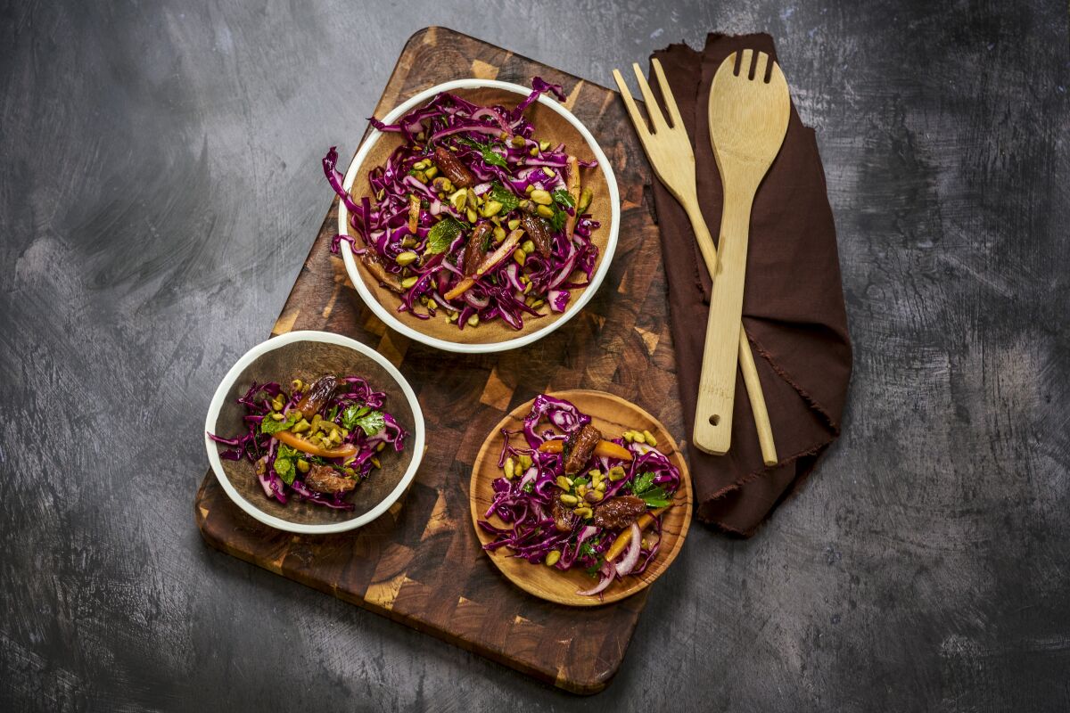 Three bowls of Red Cabbage, Date and Salted Citrus Salad on a wood cutting board, with a serving fork and spoon on a napkin