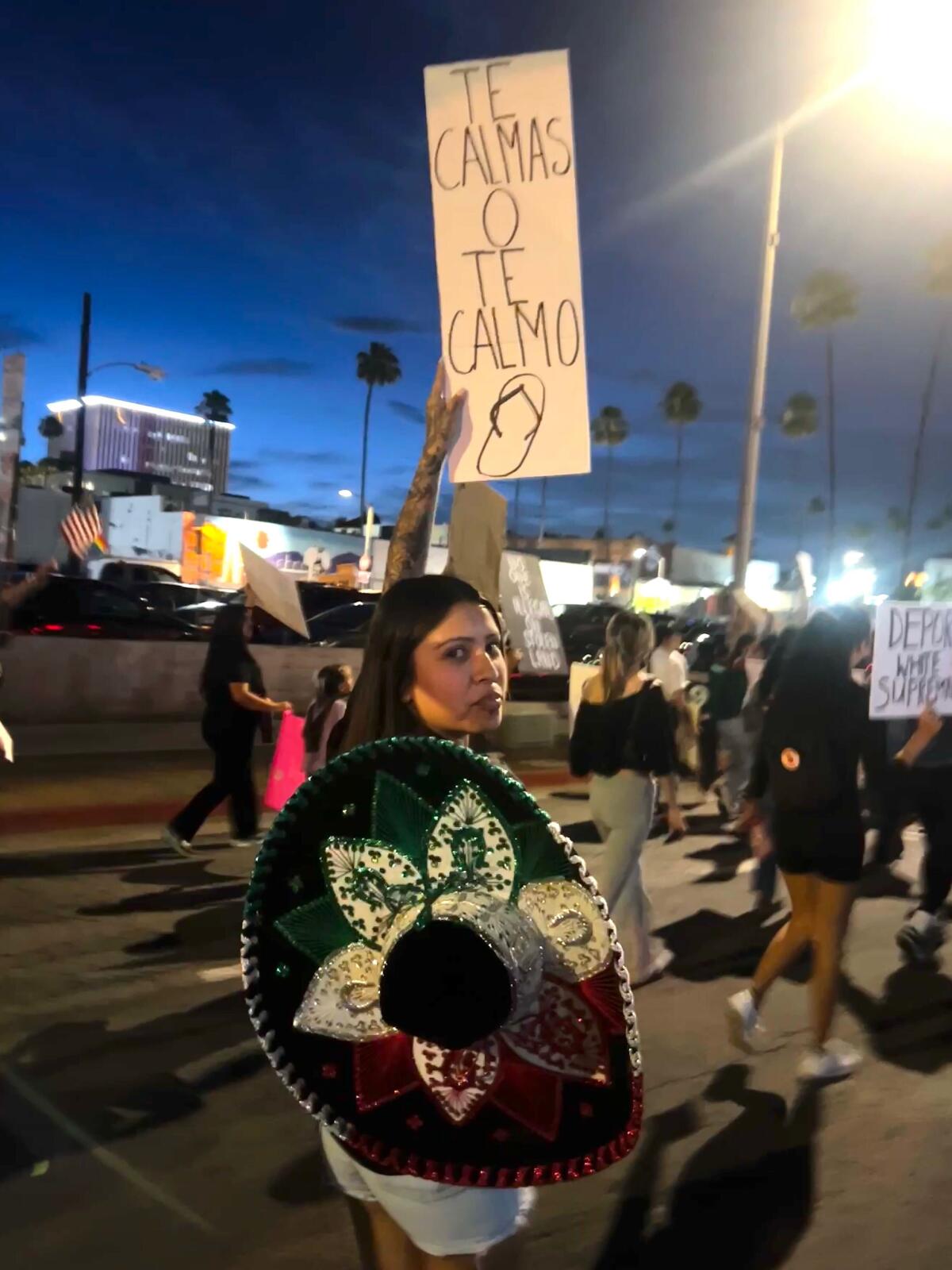 A woman stands at a protest with a person holding a sign written in Spanish.