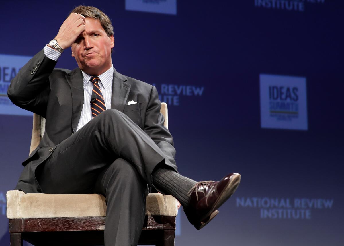 Tucker Carlson is seated on a stage, with his legs crossed, scratching his head.