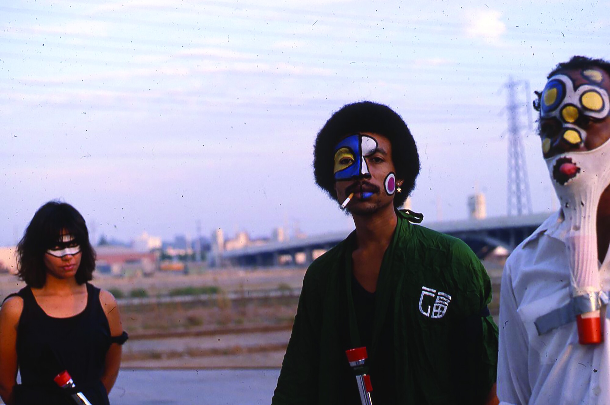 Three performers stand before a bridge in L.A.; they wear dramatic face make up and one wears a mask made from a sock