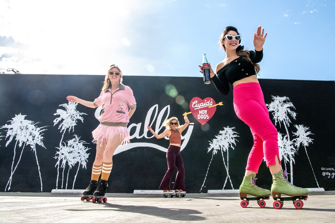 April 1: Women on roller skates in front of a wall with white images of palm trees against a black backdrop