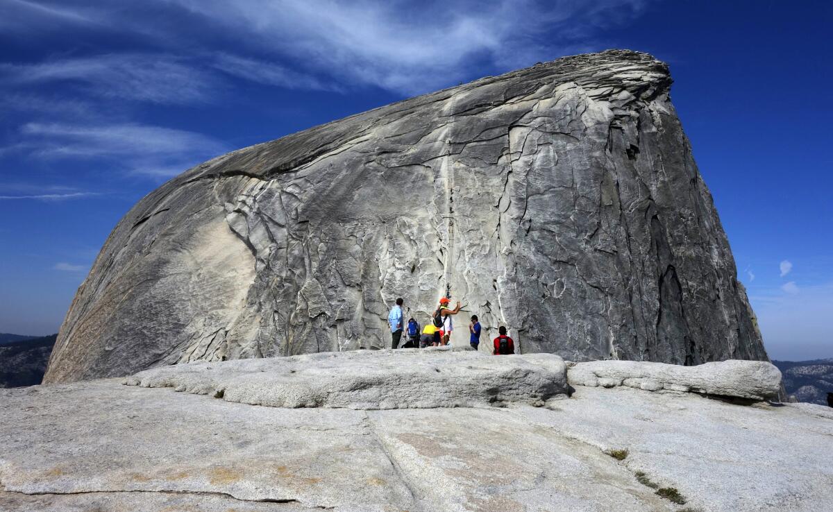 Hikers gather in the foreground as climbers use the assistance of cables to scale a granite dome