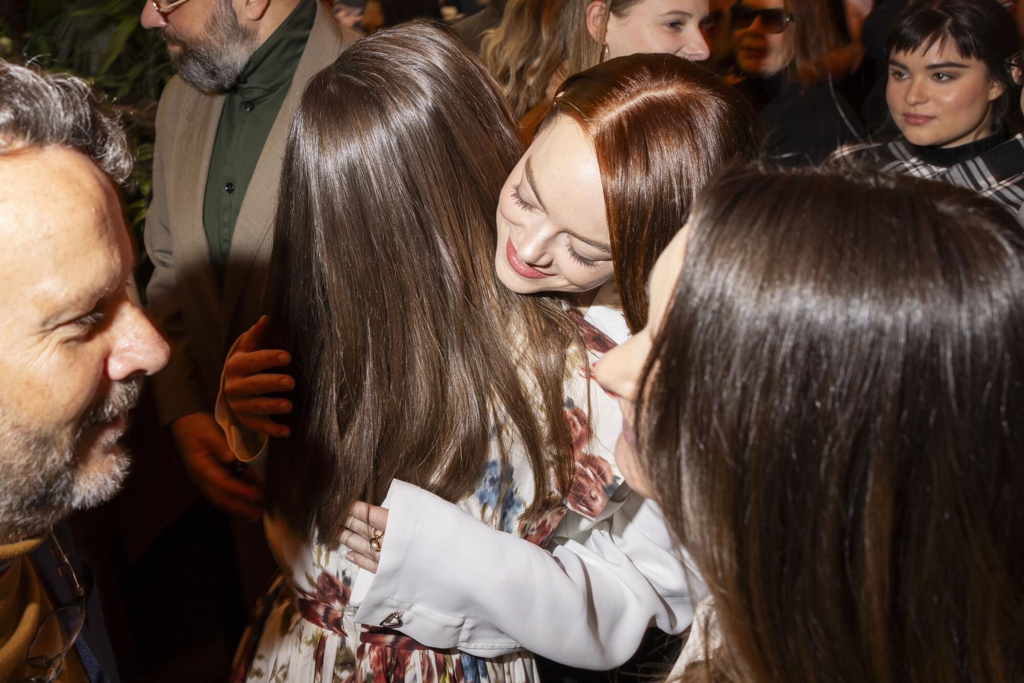 Two women with long straight hair embrace among a crowd of people.
