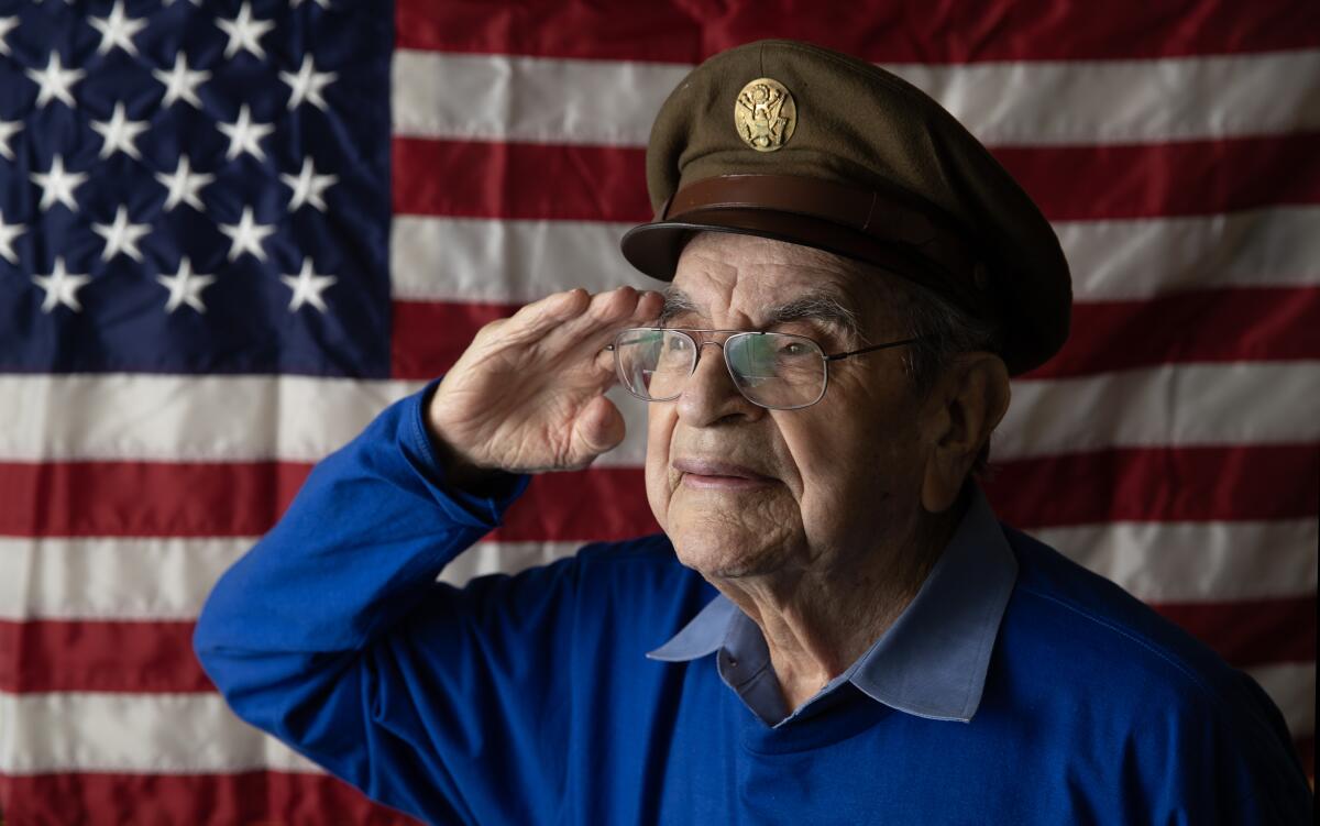 An older man wearing a brown military hat salutes in front of a U.S. flag.