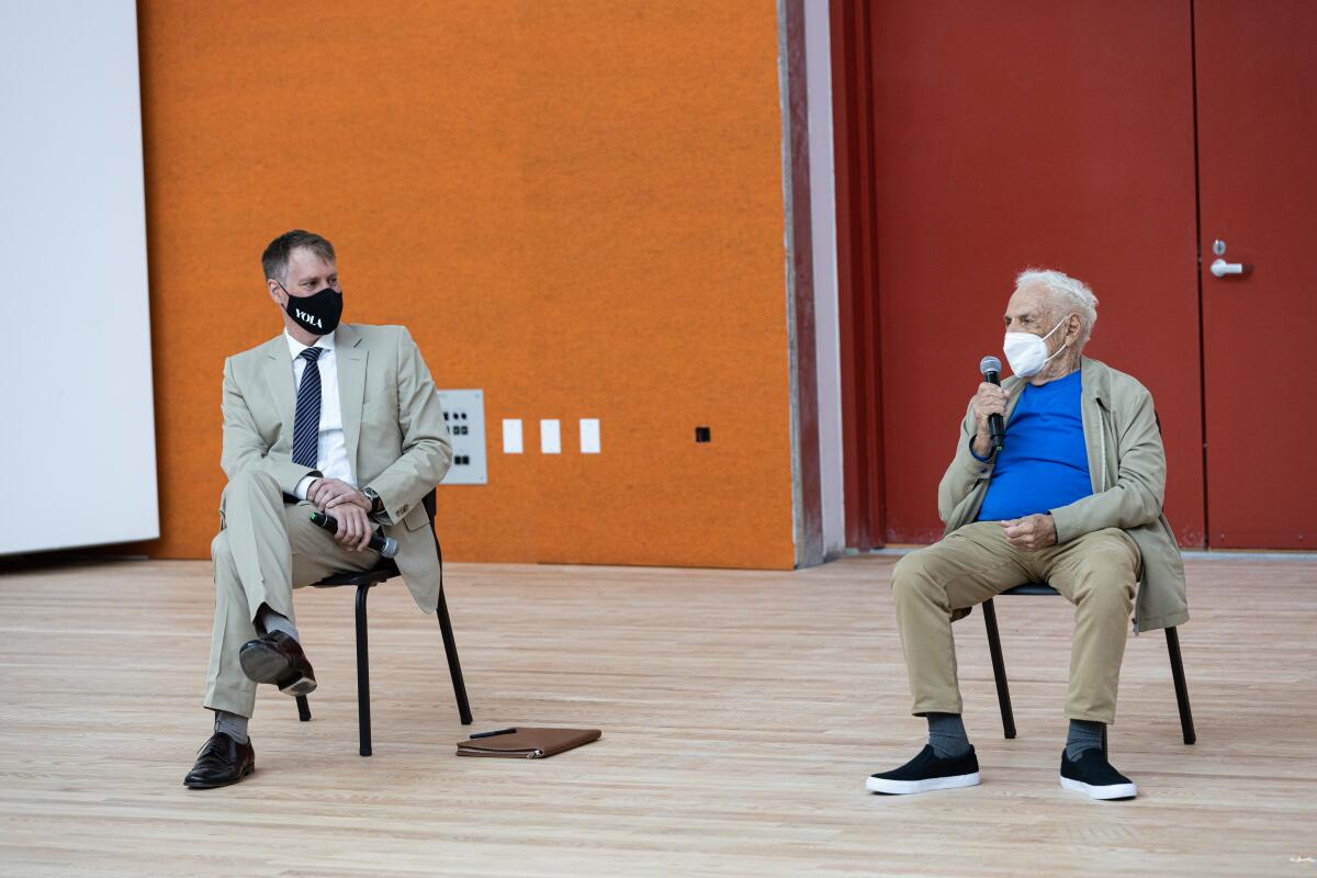 Two men, wearing masks and holding microphones, are interviewed as they sit in chairs on an empty stage.