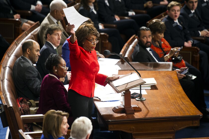 Rep. Maxine Waters stand holding up a paper while surrounded by seated men and woman.