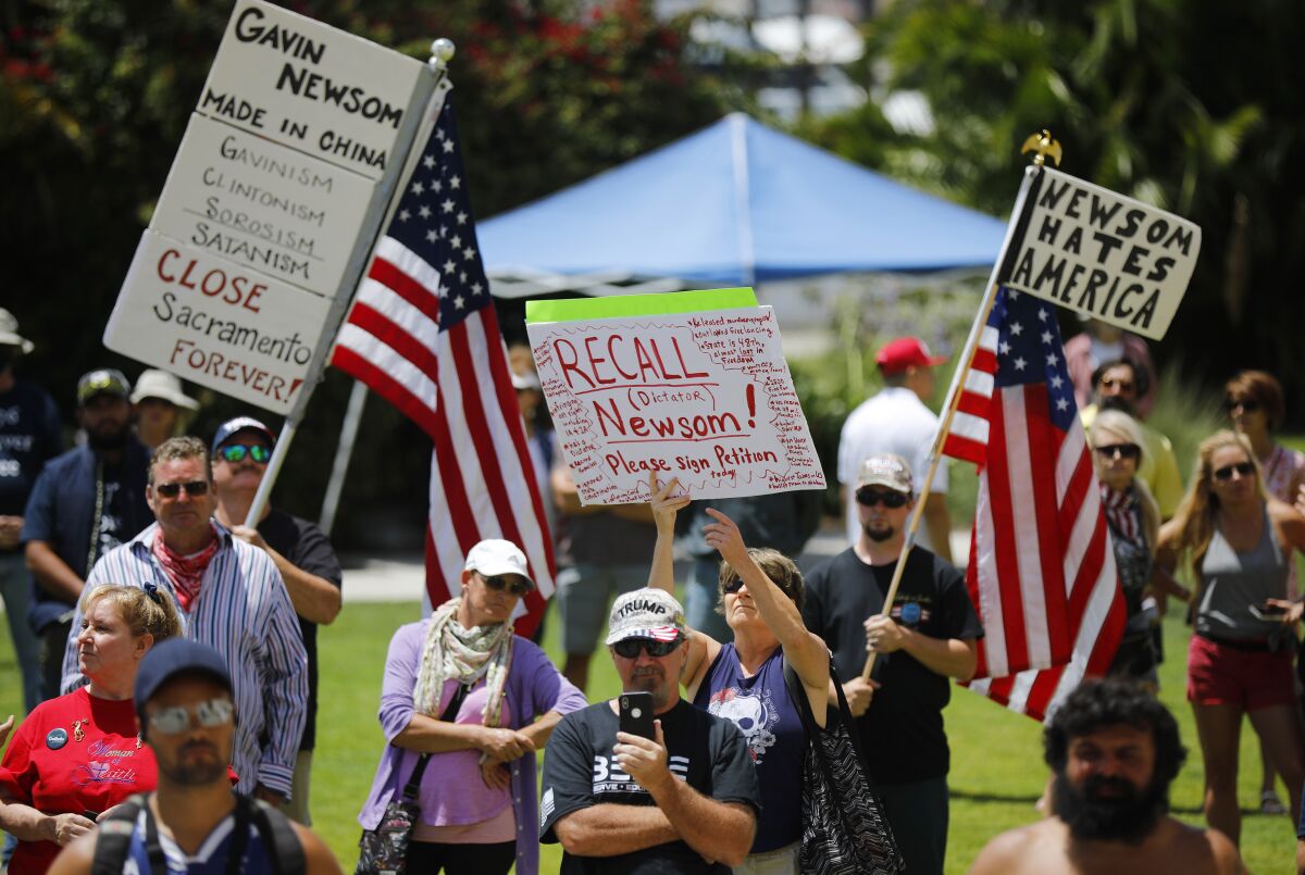 People stand outdoors in a group, holding U.S. flags and signs; one sign says "Newsom hates America."