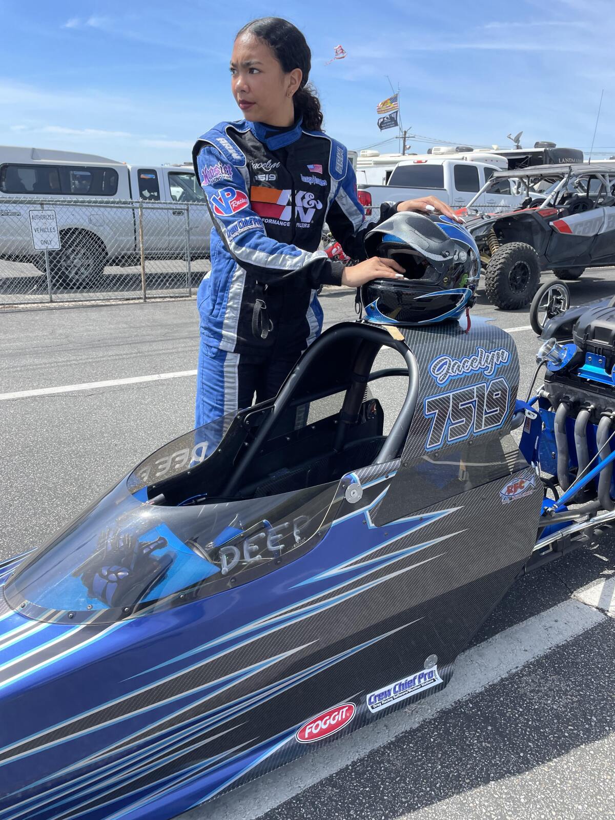 Jacelyn Gonzaga, 15, in racing suit and holding her helmet, stands beside her car at drag racing strip
