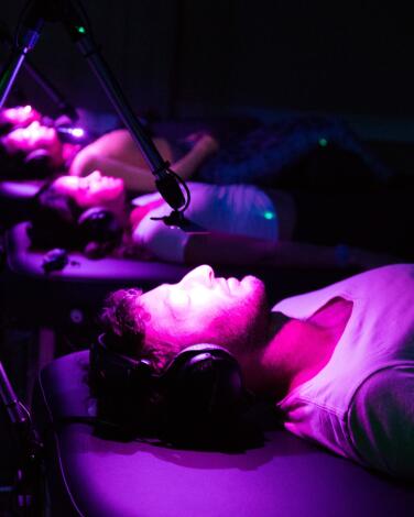 Four people on massage tables wearing headphones in a room bathed in purple light 