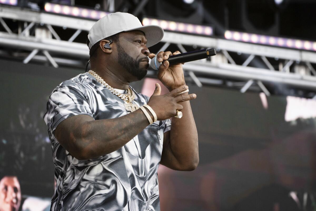 50 Cent, in a gray and white shirt and tan ball cap, raps into a microphone onstage.