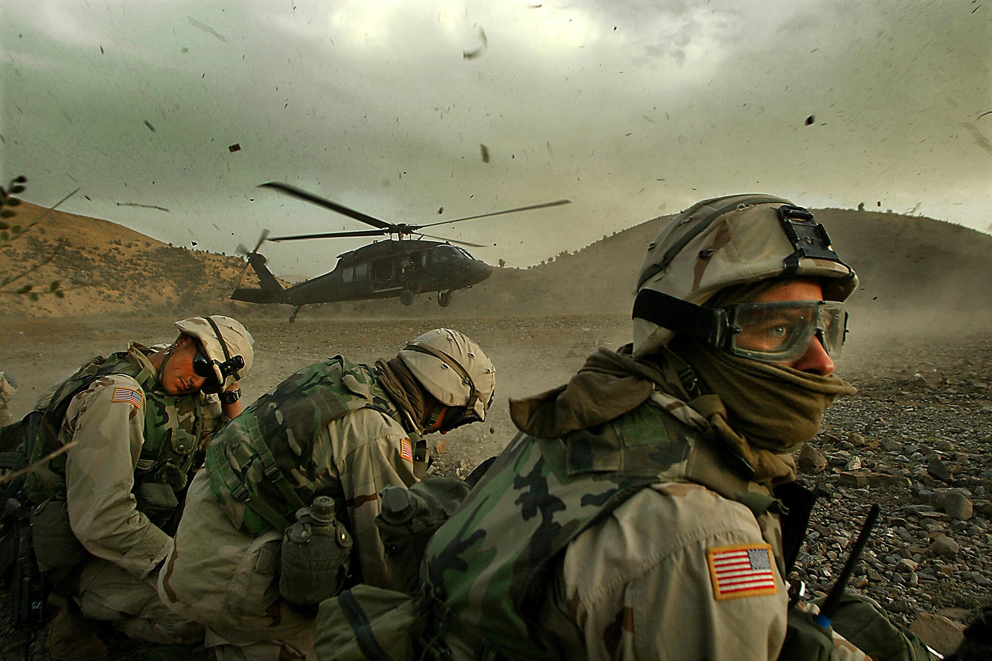 Soldiers turn their heads away from a landing helicopter kicking up dust and debris
