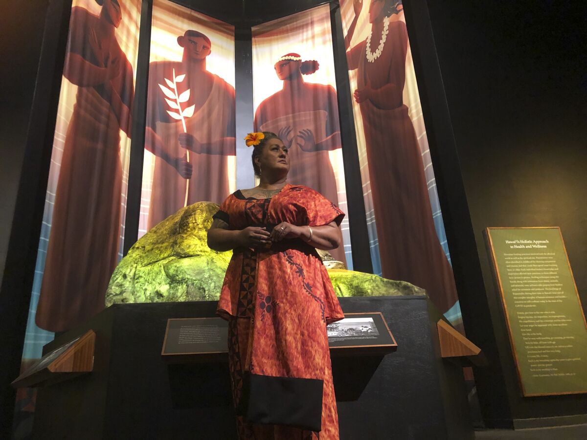 Hinaleimoana Wong-Kalu, one of the curators of the new Kapaemahu exhibit at Bishop Museum, poses for a photo in Honolulu on Thursday, June 16, 2022 in front of pictures of four healers who visited Hawaii from Tahiti more than 500 years ago. The exhibit draws attention to the stories of the healers, who were "mahu" or individuals who presented themselves as a mixture of male and female, and highlights gender fluidity's deep roots in Polynesia. (AP Photo/Audrey McAvoy)