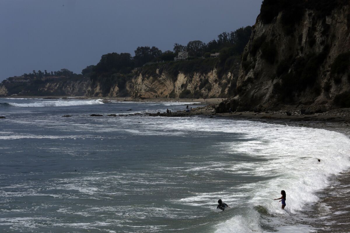 Malibu'da bir plajda çocuklar sörf yapıyor ve sağda arka planda kayalıklar görünüyor.