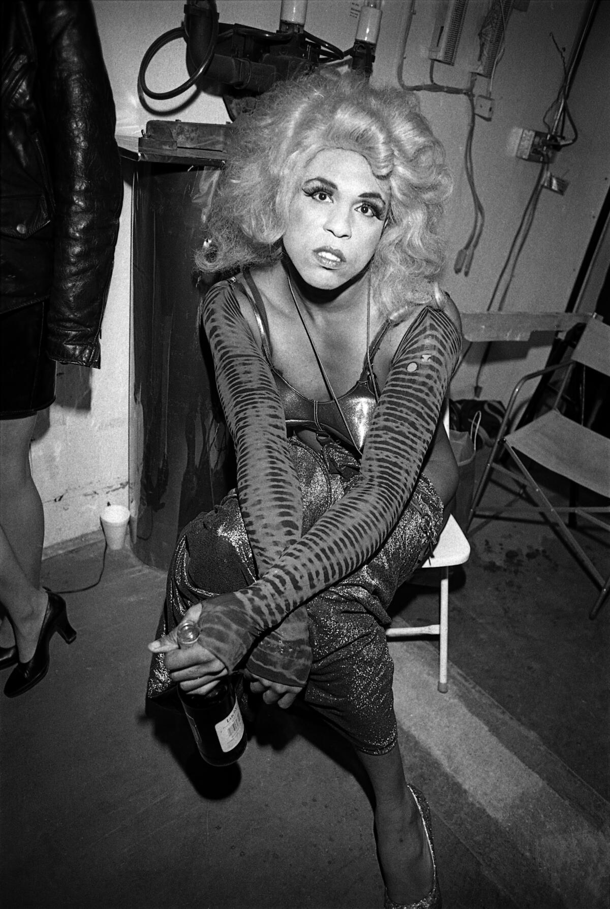 Performance artist Vaginal Davis, decked out in a blonde wig and cone bra, sits while holding a drink