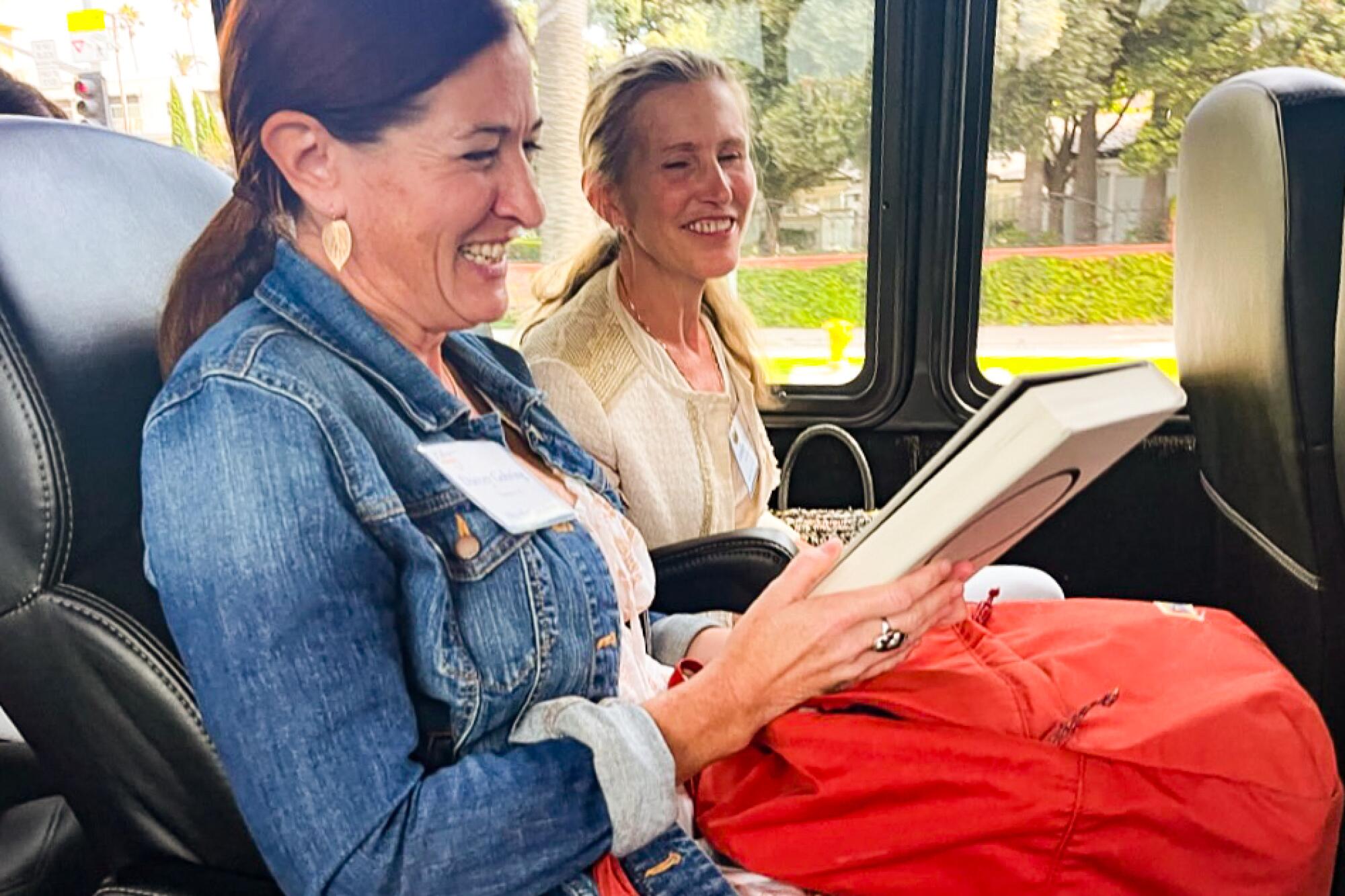 Two smiling women sit side-by-side on a bus, one holding a book