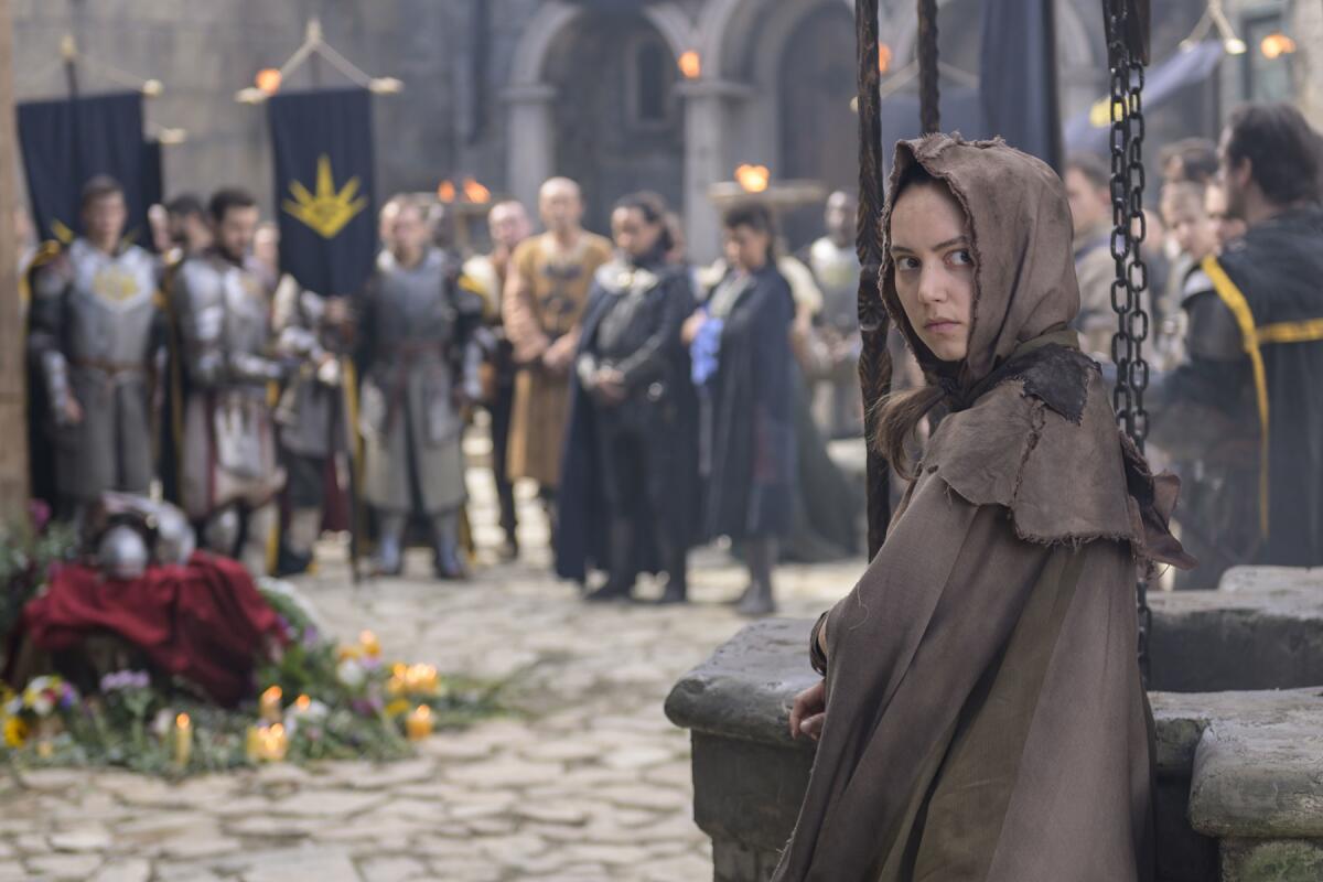 A woman in medieval garb stands in the foreground while villagers stand in the background