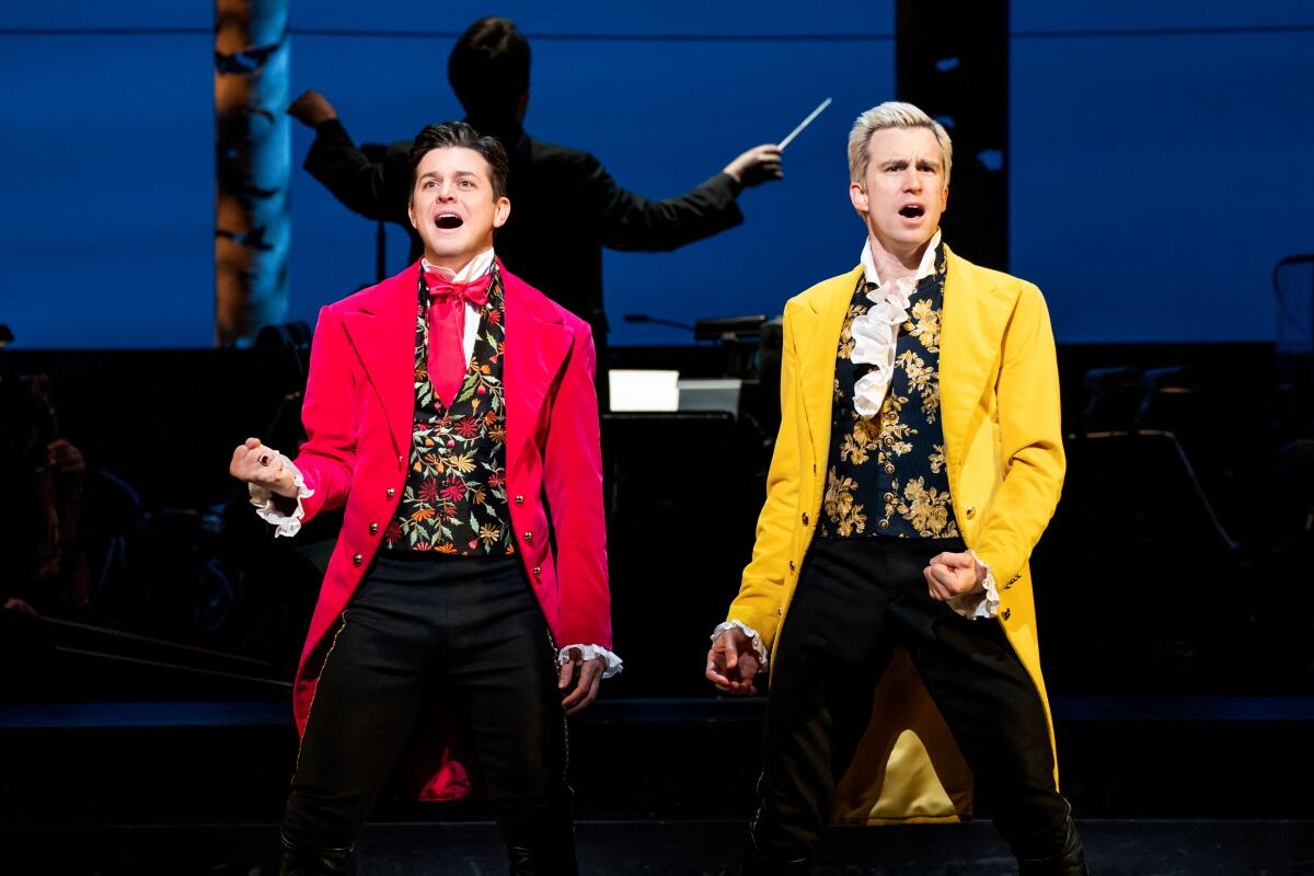 A man in a bright red jacket, red tie and flowered vest sings onstage next to a man in a similar yellow ensemble.