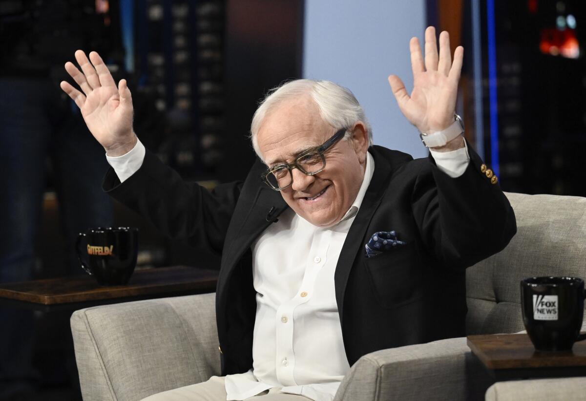 A man with white hair wearing a suit, sitting in an armchair and raising his hands into the air