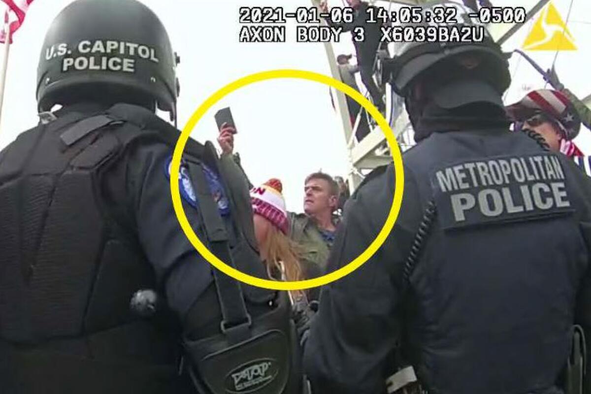 Police officers engage in a standoff with a crowd. A man in a green jacket holding up a cell phone is circled in yellow.