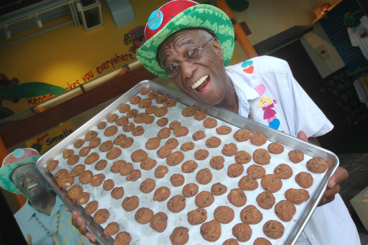 Wally Amos, in a colorful hat, smiles while holding a tray of cookies