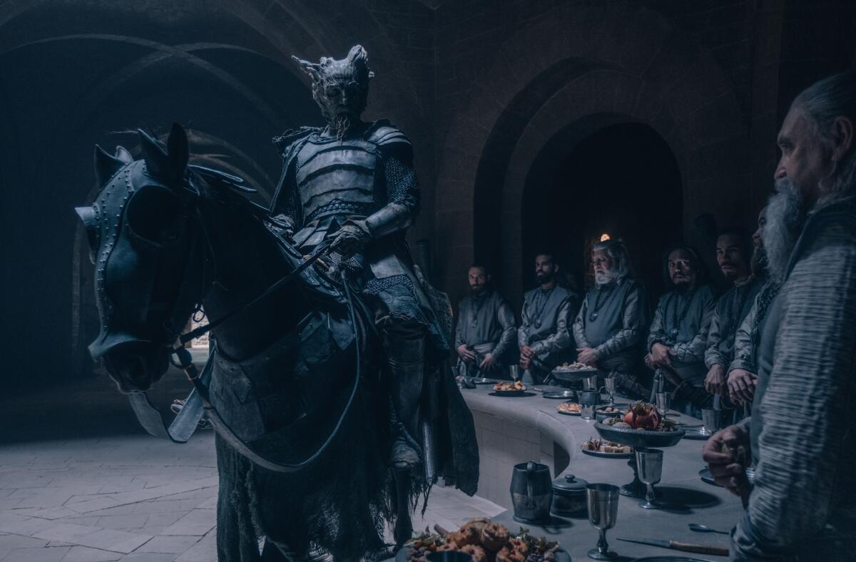 Ralph Ineson in armor astride a horse in front of a table behind which stand several people in "The Green Knight."