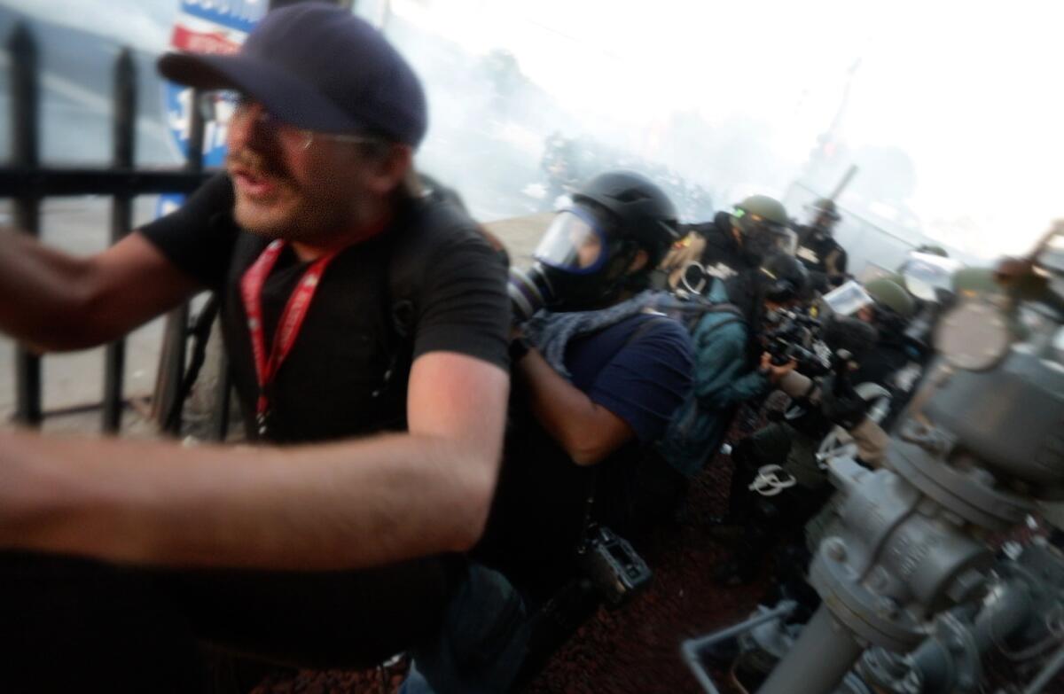 A man with press credentials around his neck runs from law enforcement in gas masks and riot gear.