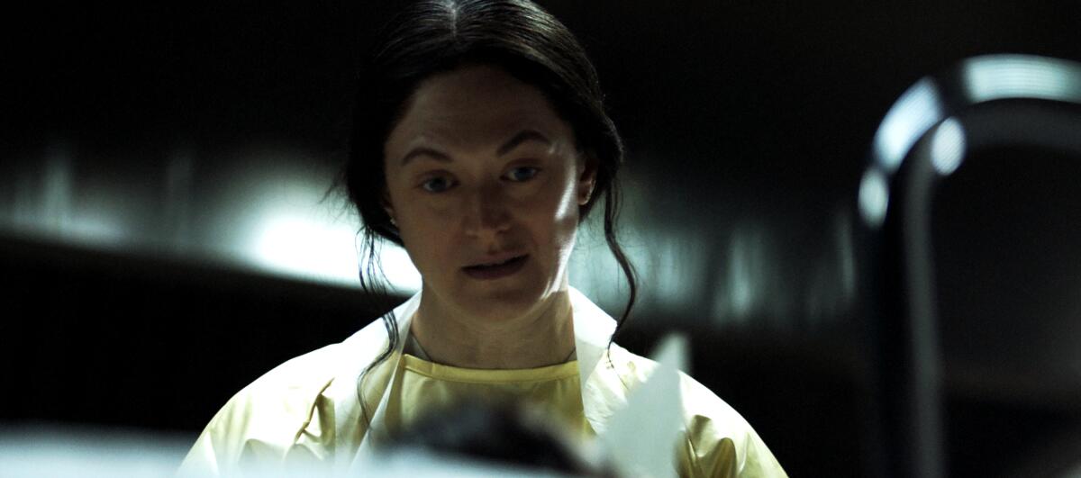 A woman in a yellow surgical smock stares at an offscreen corpse.