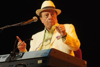 Sergio Mendes in a white suit and a hat holding up his hands while he sits at a keyboard