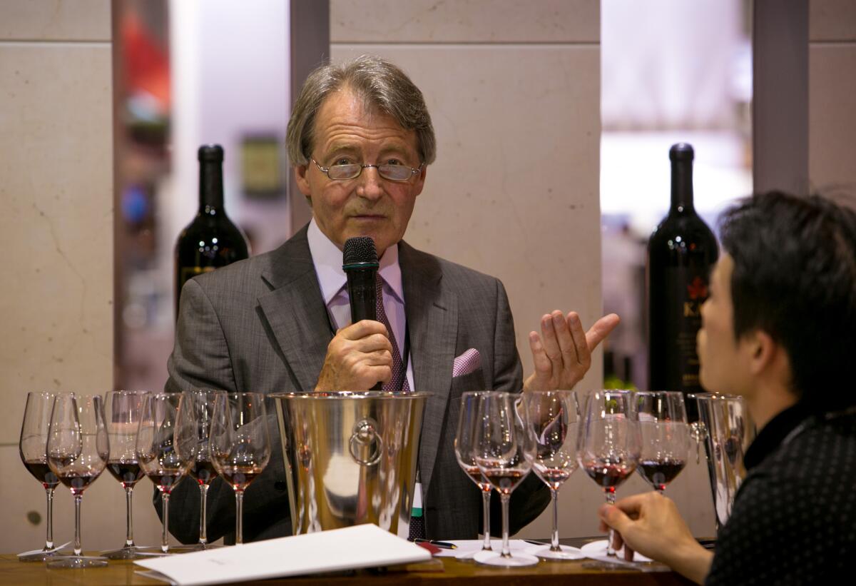 A man in a suit and tie speaks into a microphone behind several wine glasses lined up for a tasting