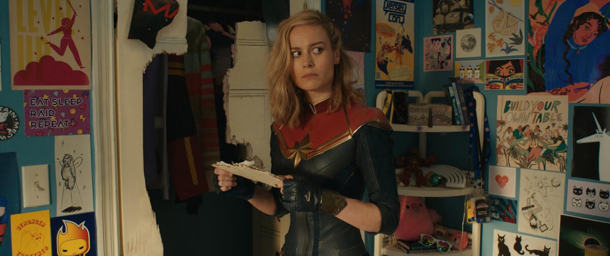 Brie Larson holds a piece of paper in a room covered in posters. She wears a Captain Marvel costume