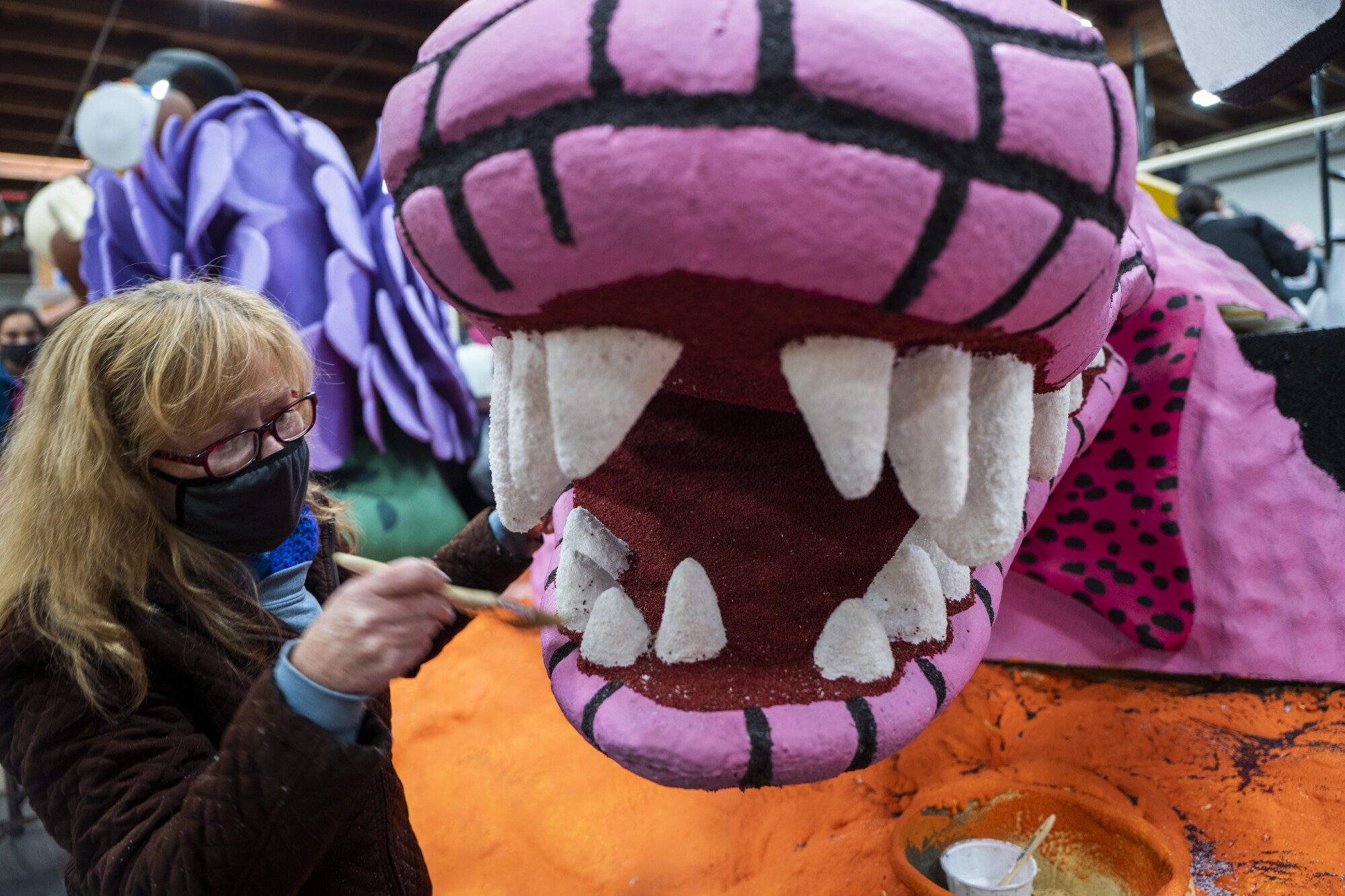 A masked woman paints a creature on a Rose Parade float.