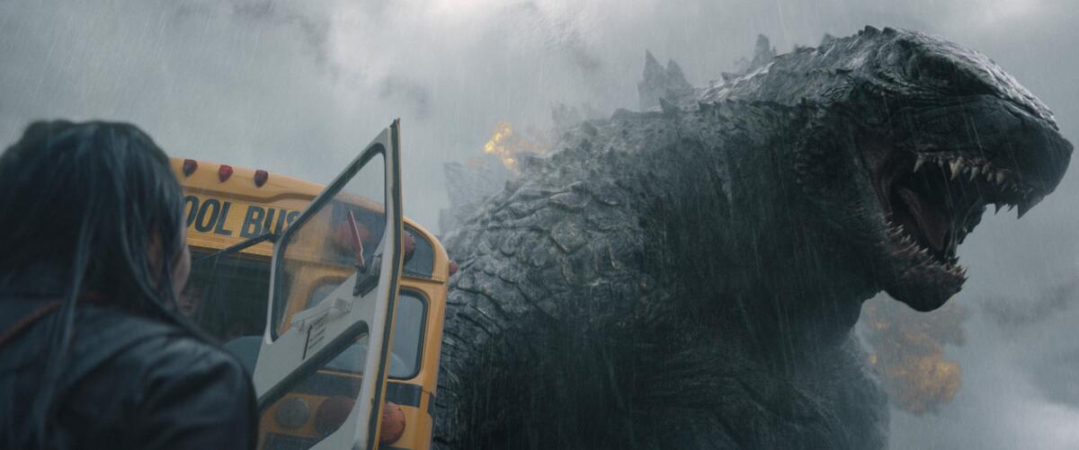 Godzilla roars from behind a crashed yellow school bus as smoke drifts around.