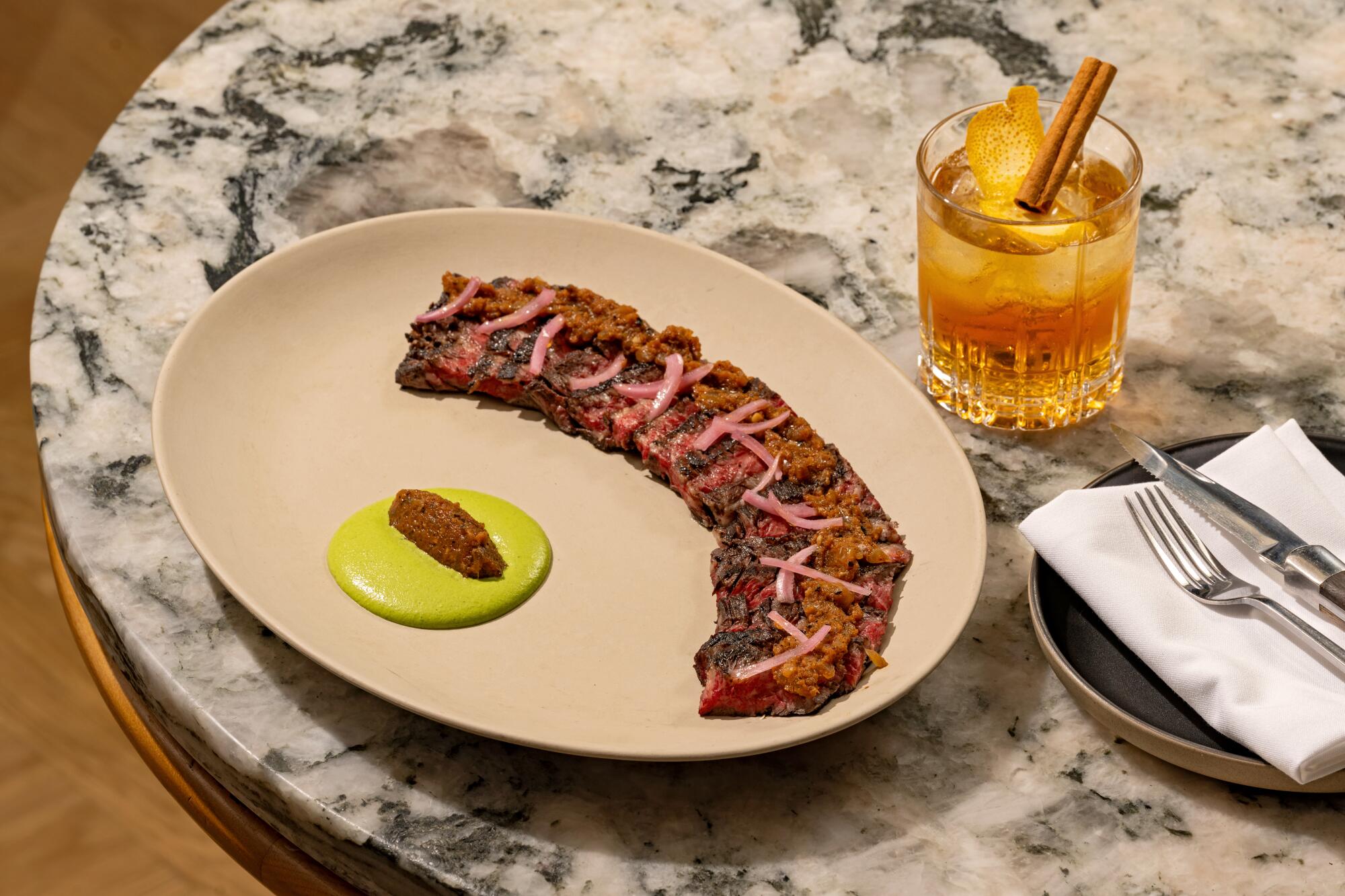 A plate of thin-sliced skirt steak with green tahini sauce, sitting on a marble tabletop next to an orange cocktail.
