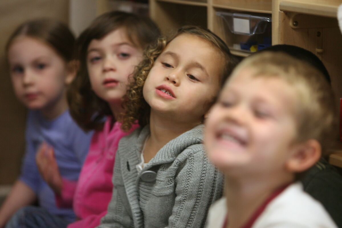 Four children sit in a row, listening to their pre-kindergarten teacher.
